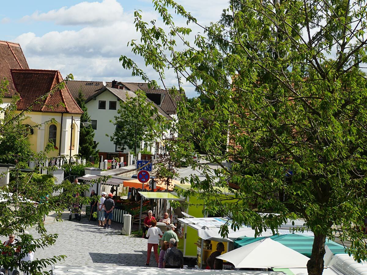 Blick auf einen belebten Dorfmarkt mit Menschen, Marktständen, alten Häusern und einer gelben Kirche unter blauem Himmel.