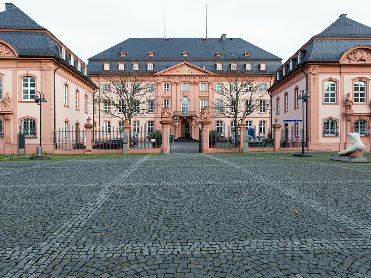 Landtag Rheinland-Pfalz in Mainz, umgeben von rosafarbenen Gebäuden, einem gepflasterten Vorplatz und einer weißen Fußskulptur.