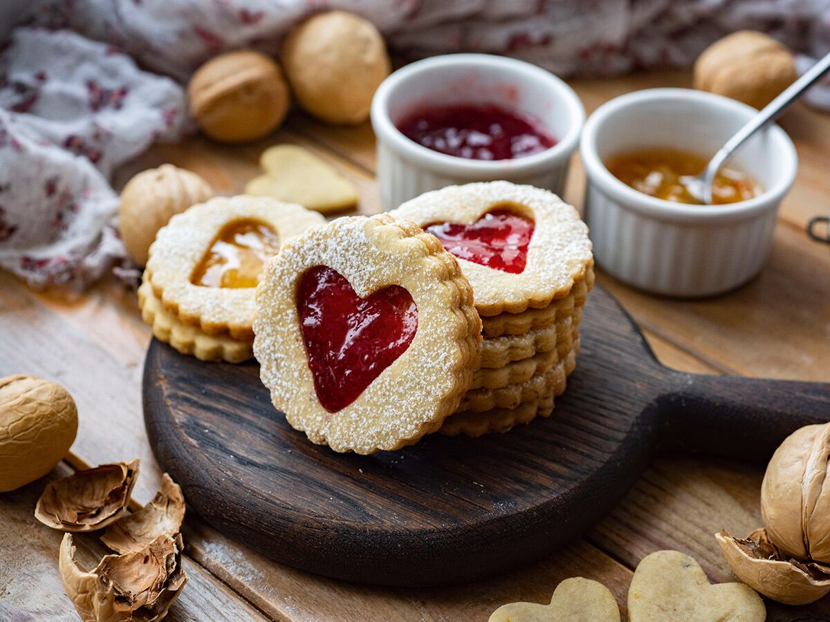 Nahaufnahme von Herz-Keksen mit roter und gelber Marmeladenfüllung auf rustikalem Holzbrett, umgeben von Walnüssen und Marmeladen.