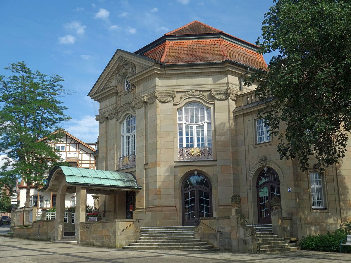 Großes, achteckiges Sandsteingebäude mit roten Ziegeln und verzierten Fenstern, umgeben von Bäumen und blauem Himmel.
