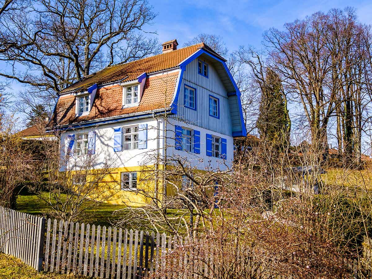 Weißes Haus mit gelbem Sockel, rotem Dach, blauen Fensterläden, Holzzaun und kahlen Bäumen unter blauem Winterhimmel.