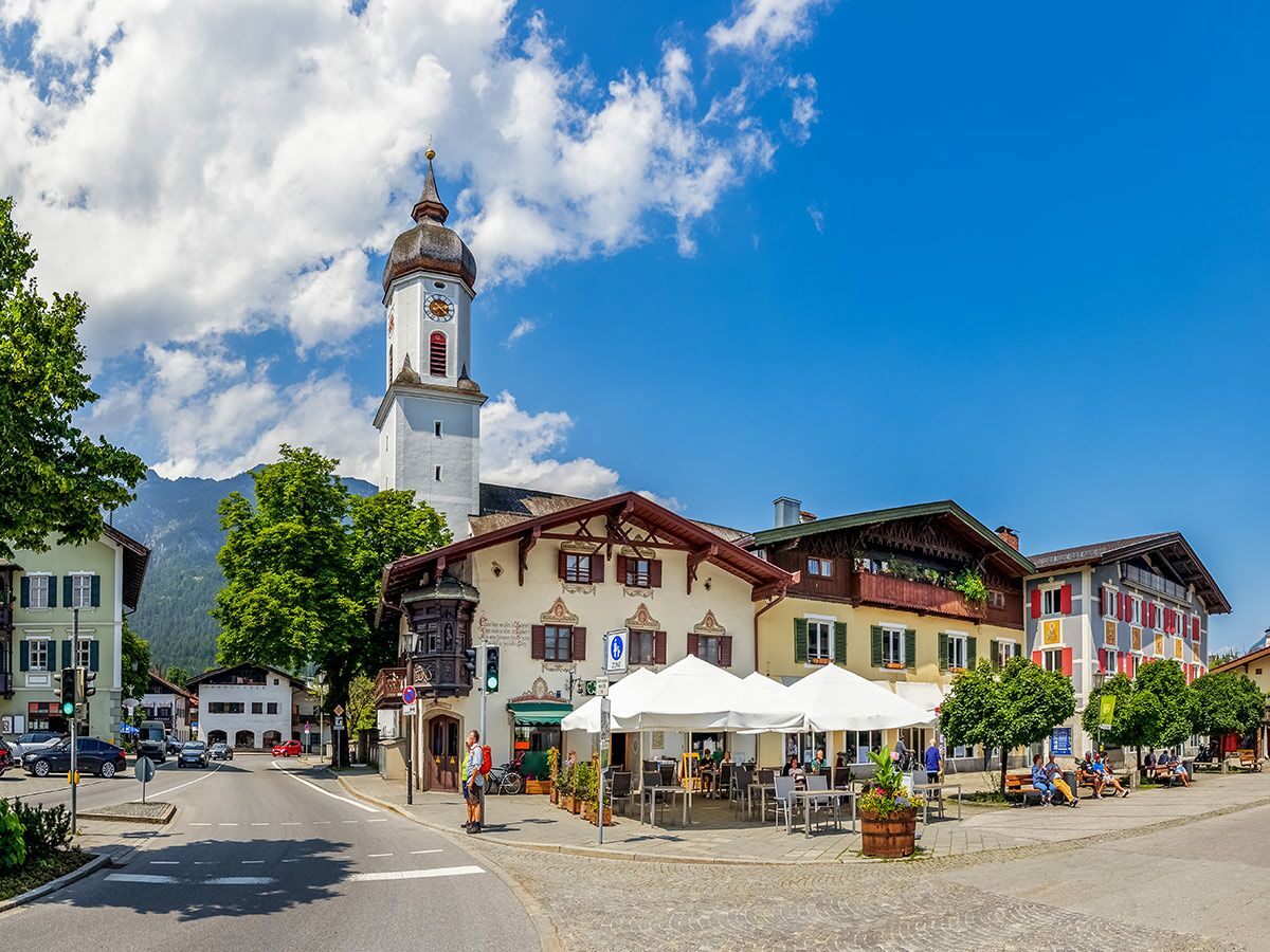 Idyllische Alpenstadt mit weißer Kirche, traditionellen Häusern, Straßencafé, Bergen und blauem Himmel an einem sonnigen Tag.