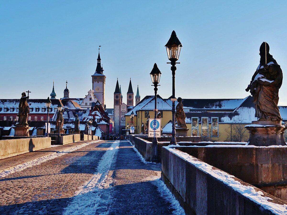 Alte Mainbrücke Würzburg im Winter, bedeckt mit Schnee, Statuen und historischen Türmen im Hintergrund bei sonnigem, blauem Himmel.