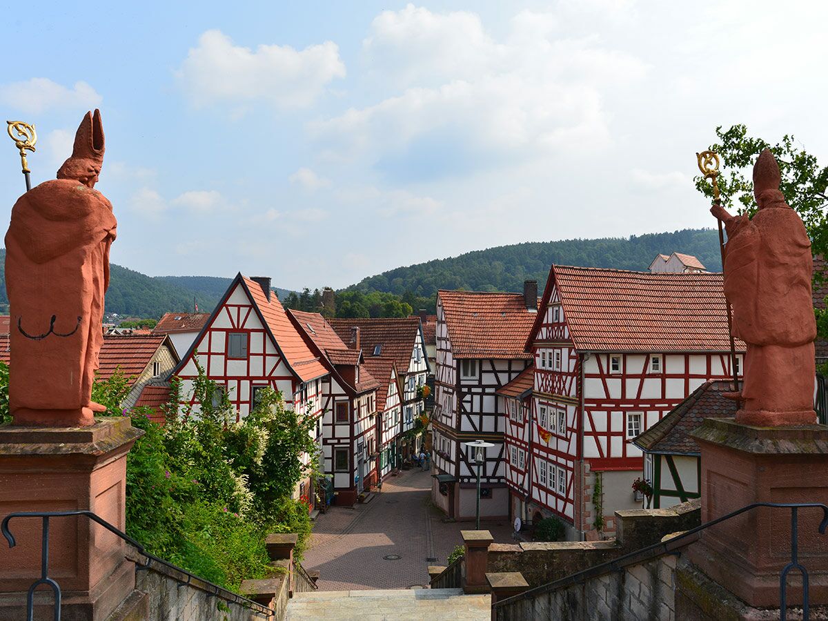 Blick von einer Treppe auf ein malerisches deutsches Dorf mit traditionellen Fachwerkhäusern und roten Dächern, umrahmt von zwei Bischofs-Statuen.