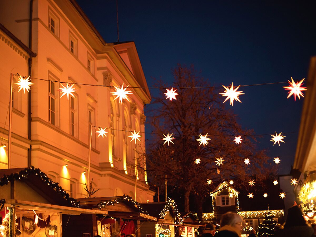 Abendlicher Weihnachtsmarkt mit leuchtenden Sternen, festlich beleuchteten Gebäuden, Marktständen und Winterbäumen.