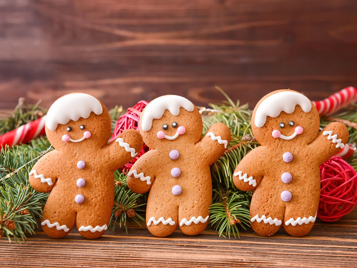 Drei Lebkuchenmänner, festlich dekoriert mit Tannenzweigen, Zuckerstange und roten Kugeln auf Holztisch.