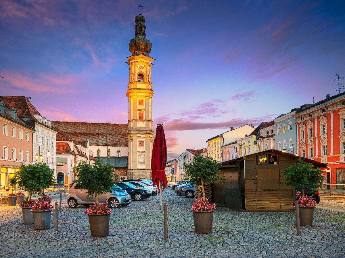 Historischer Stadtplatz bei Dämmerung mit imposantem Kirchenturm, farbenfrohen Gebäuden, Kopfsteinpflaster und geparkten PKWs.