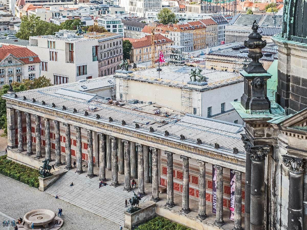 Luftaufnahme des Alten Museums in Berlin mit Säulen, Treppe, Brunnen und Menschen im Vordergrund, umgeben von Stadtgebäuden.
