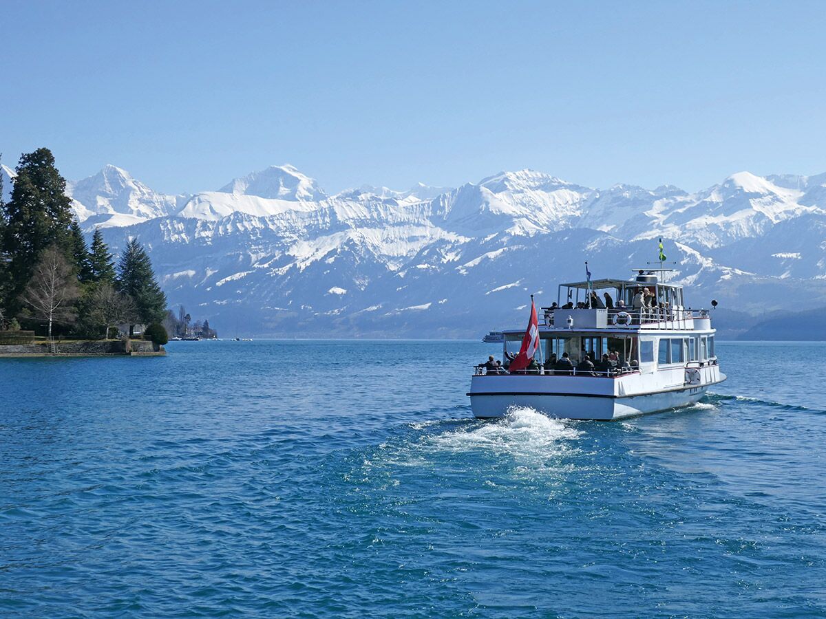 Ein weißes Passagierboot fährt auf einem blauen Schweizer See. Im Hintergrund erheben sich schneebedeckte Alpen und grüne Ufer unter blauem Himmel.