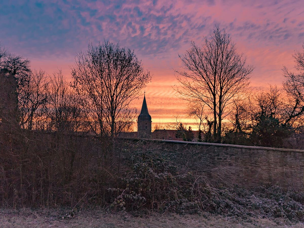 Dramatischer Sonnenuntergang mit pink-orangefarbenem Himmel über einem Kirchturm und kahlen Bäumen im winterlichen Frost.