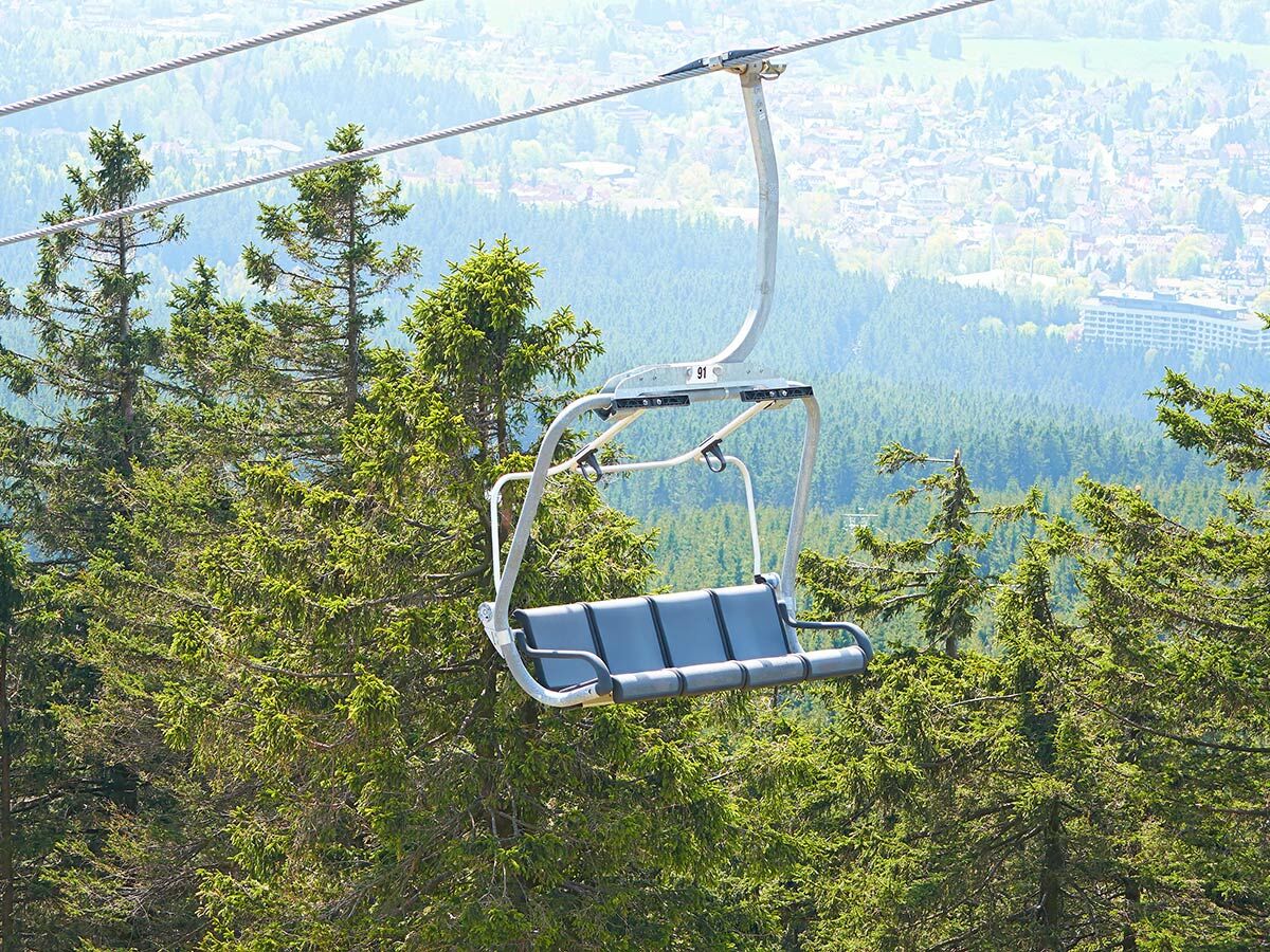 Leerer Sessellift schwebt über dichten grünen Wald mit einer Stadt und dunstigem Himmel im Hintergrund.