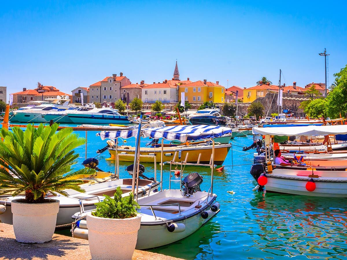 Blick auf den belebten Hafen von Budva, Montenegro, mit zahlreichen Booten, Yachten und bunten Gebäuden unter strahlend blauem Himmel.
