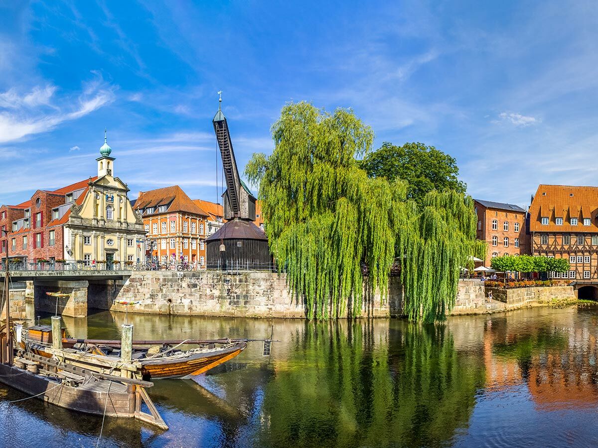 Panoramablick auf Lüneburgs Alter Kran, historische Gebäude und Weiden am Flussufer unter blauem Himmel mit Booten.