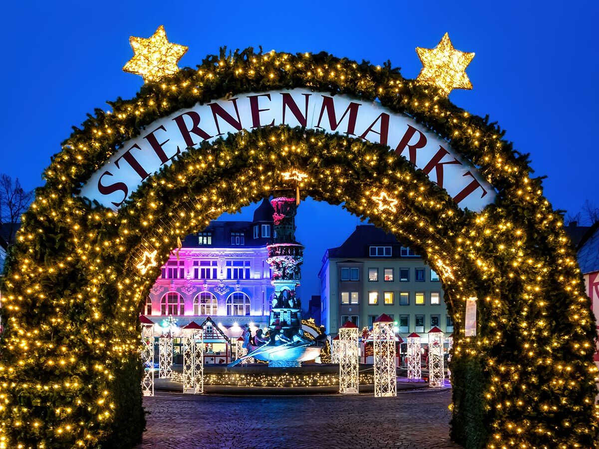 Festlich beleuchteter Eingangsbogen zum Sternenmarkt mit goldenen Lichtern und Sternen bei Dämmerung. Im Hintergrund ein beleuchteter Brunnen und Gebäude.