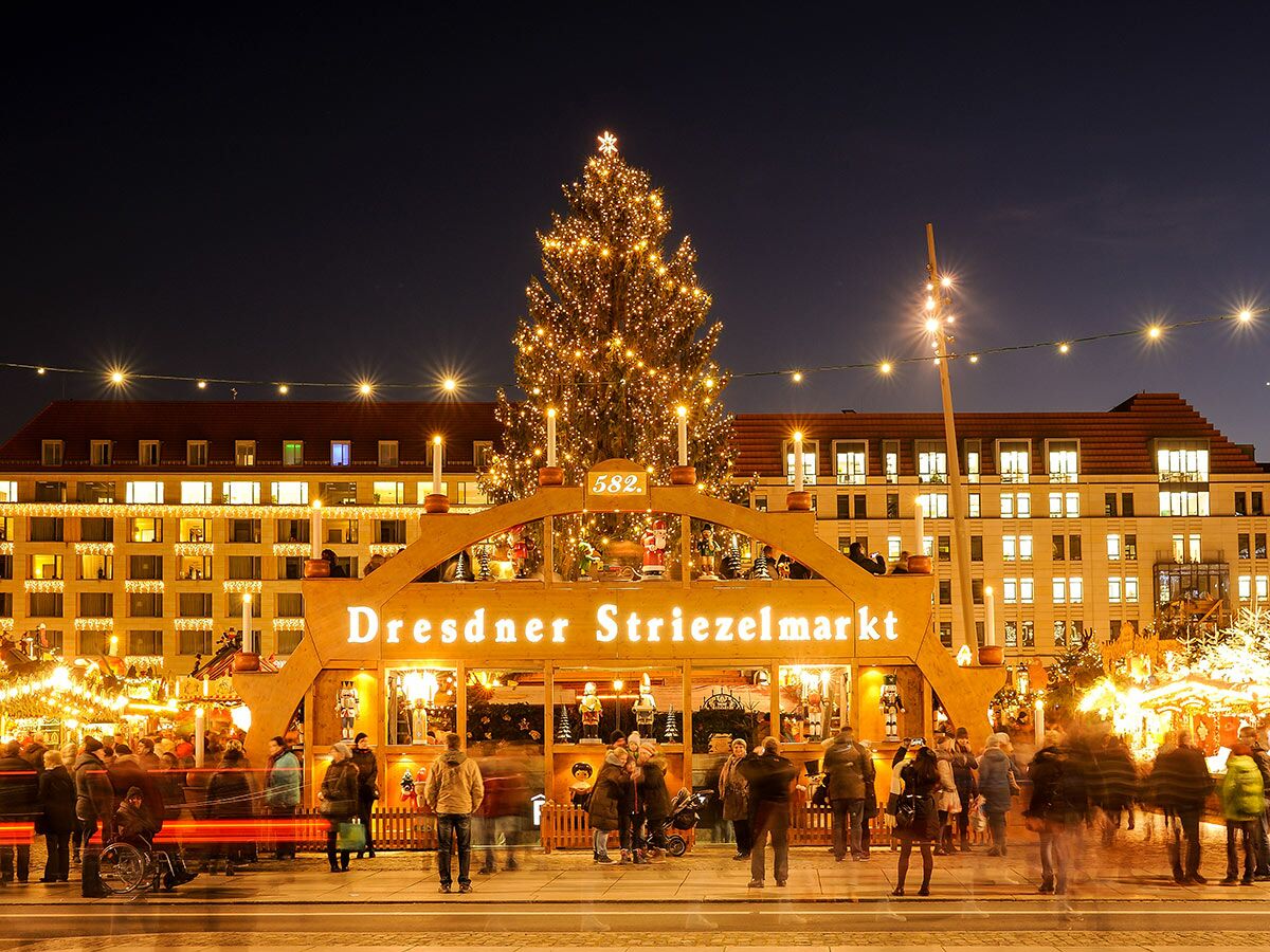 Abendaufnahme des festlichen Dresdner Striezelmarktes mit beleuchtetem Weihnachtsbaum, Holzhütten und vielen Besuchern.