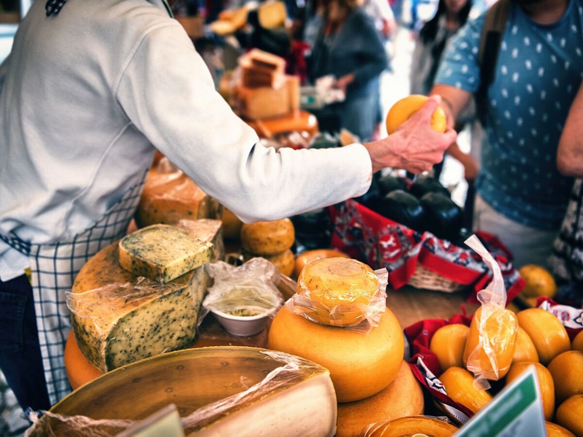 Marktverkäufer im karierten Kittel reicht auf einem geschäftigen Käsemarkt ein Produkt an einen Kunden.