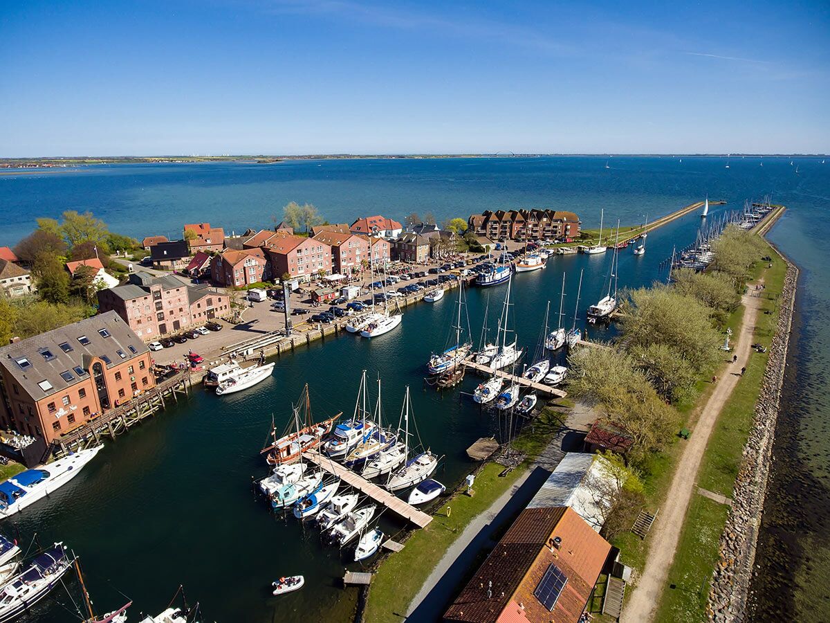 Luftaufnahme eines malerischen Yachthafens in einer Küstenstadt mit vielen Segelbooten und historischen Gebäuden am Wasser unter blauem Himmel.