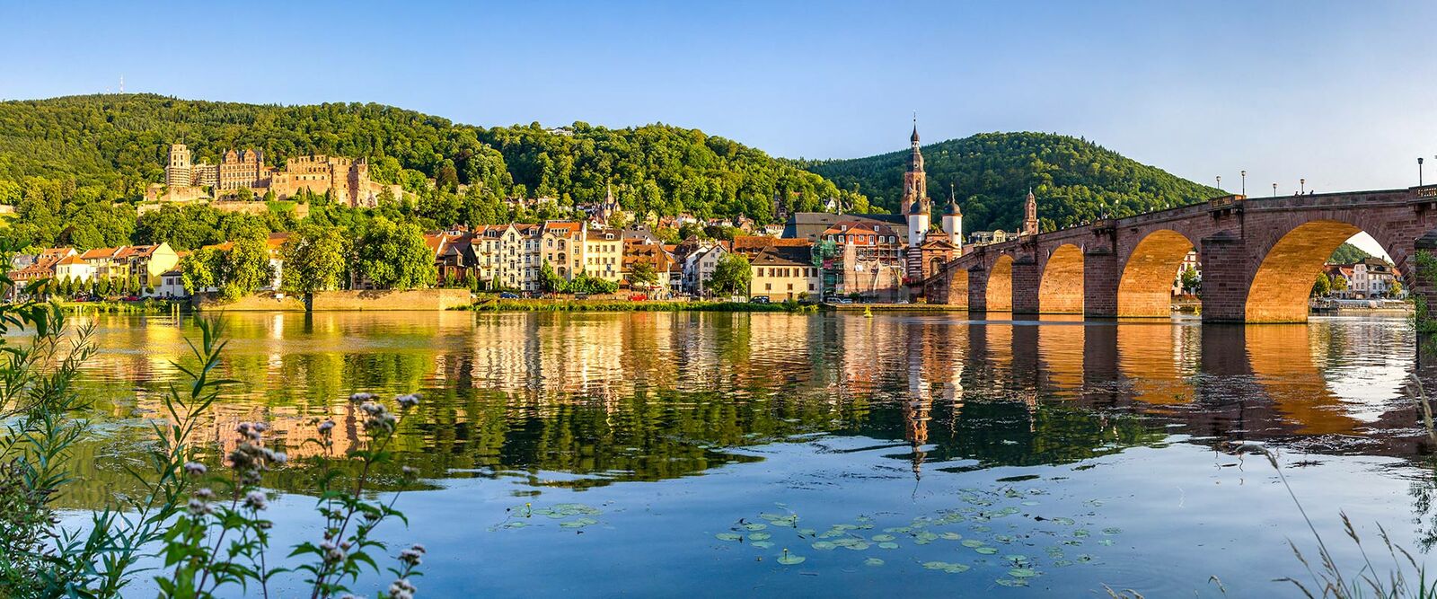 Panoramablick auf Heidelberg mit Schloss, Altstadt, Karl-Theodor-Brücke und Neckarfluss bei Sonnenuntergang, grüne Hügel und Reflexionen.