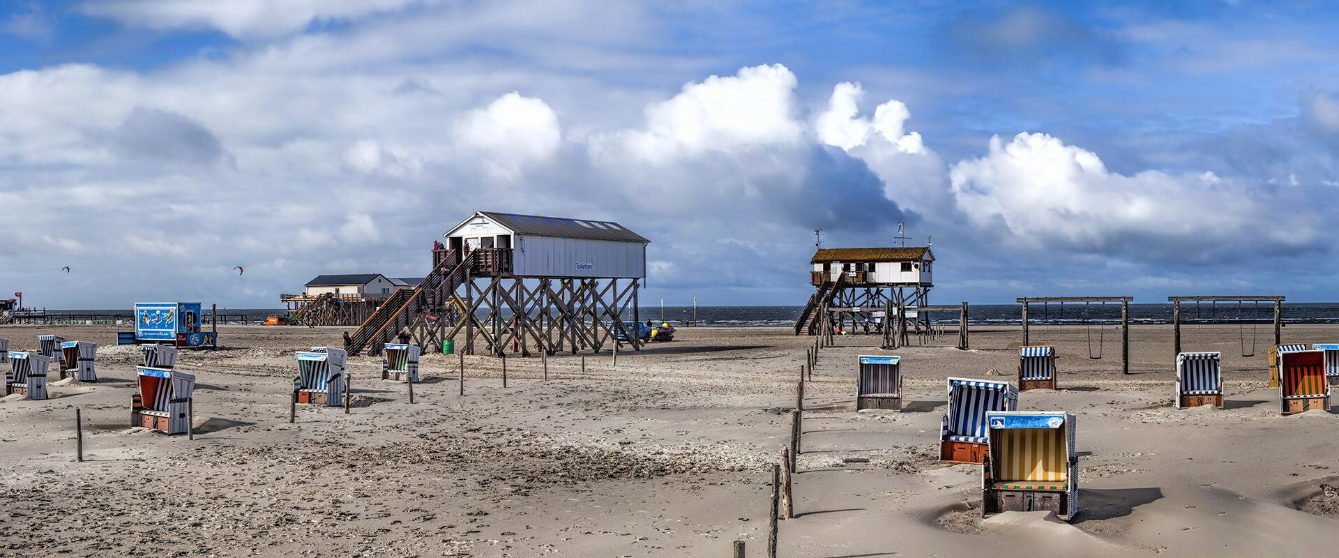Panoramabild eines Nordseestrands mit Pfahlbauten, zahlreichen gestreiften Strandkörben, Schaukeln und Kitesurfern unter weitem Wolkenhimmel.
