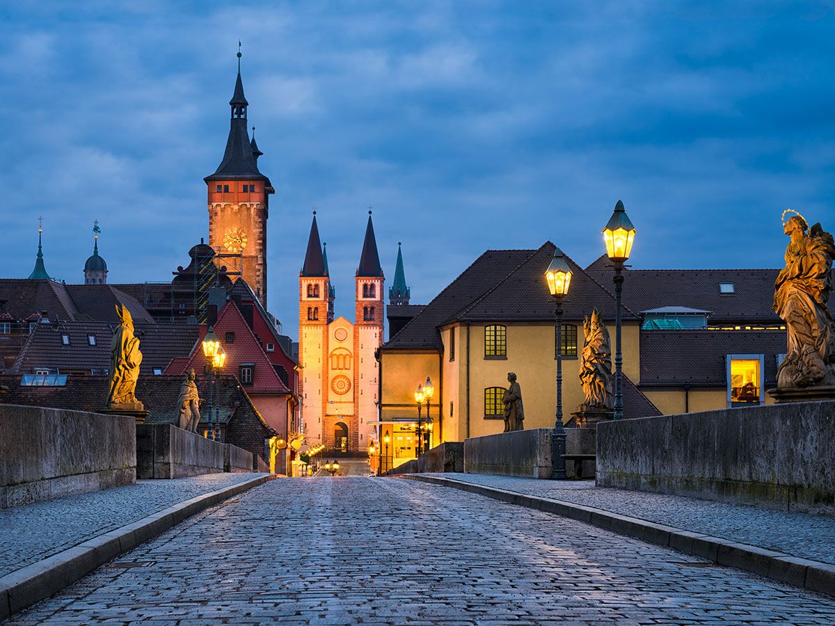 Alte Mainbrücke in Würzburg bei Dämmerung mit historischen Statuen, beleuchteten Laternen und Stadtkulisse mit Kirchen.