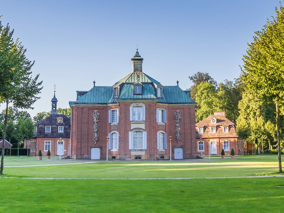 Panoramablick auf ein historisches Backsteinschloss mit grüner Dachkuppel und hellblauen Fensterläden, umgeben von gepflegten Rasenflächen und Bäumen unter blauem Himmel.