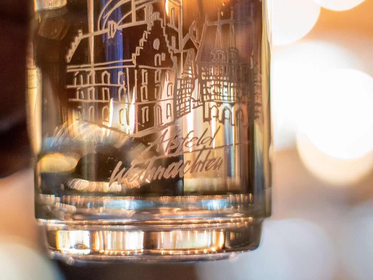 Nahaufnahme eines Glases mit Gravur von Alsfelder Gebäuden und dem Schriftzug 'Alsfeld Weihnachten' vor warmen Bokeh-Lichtern.