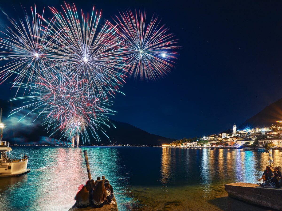 Spektakuläres Feuerwerk in Rot, Blau und Weiß über einem beleuchteten Dorf am See, Menschen beobachten das Spektakel bei Nacht.