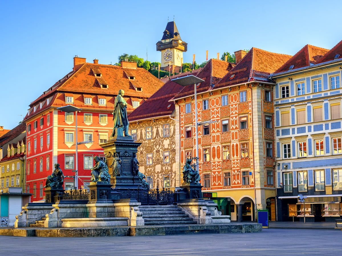 Historisches Stadtbild von Graz, Österreich, mit bunten Gebäuden, Denkmal und Uhrturm am Schlossberg unter blauem Himmel.