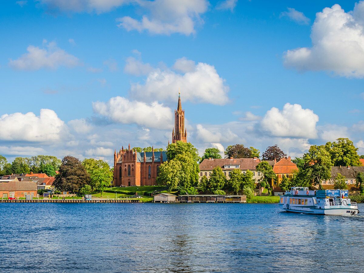 Blick über einen blauen See auf eine rote Backsteinkirche mit hohem Turm, umgeben von Bäumen und Häusern, mit einem Passagierschiff auf dem Wasser.