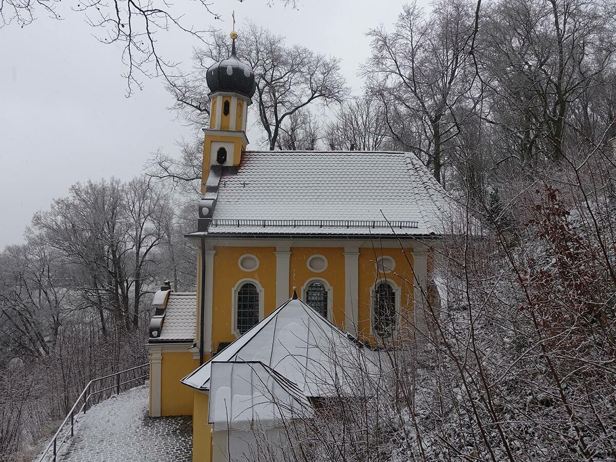 Gelbe Kapelle mit schwarzem Zwiebelturm und Schnee auf dem Dach, umgeben von schneebedeckten Bäumen im Winter.