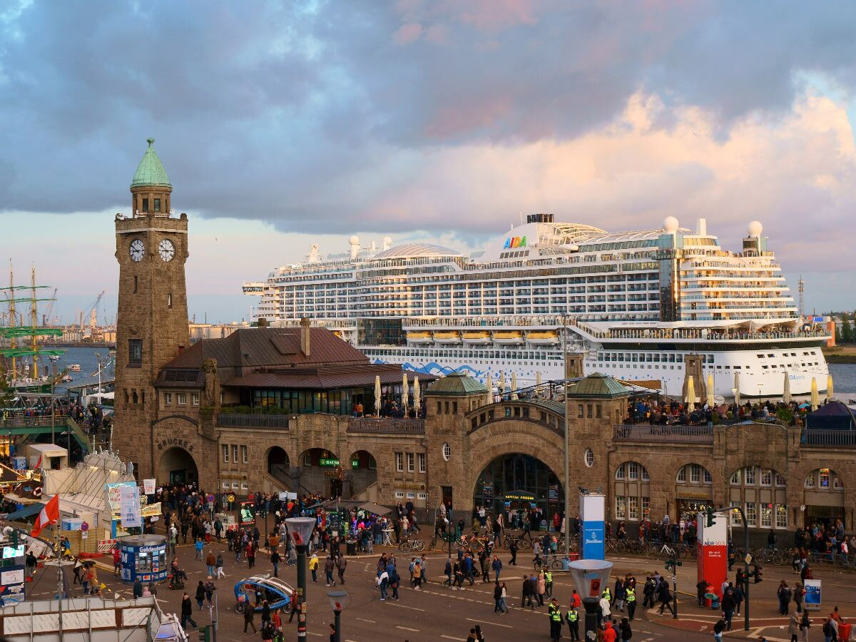Abendstimmung an den Hamburger Landungsbrücken mit Pegelturm, belebtem Boulevard und großem AIDA-Kreuzfahrtschiff im Hafen.