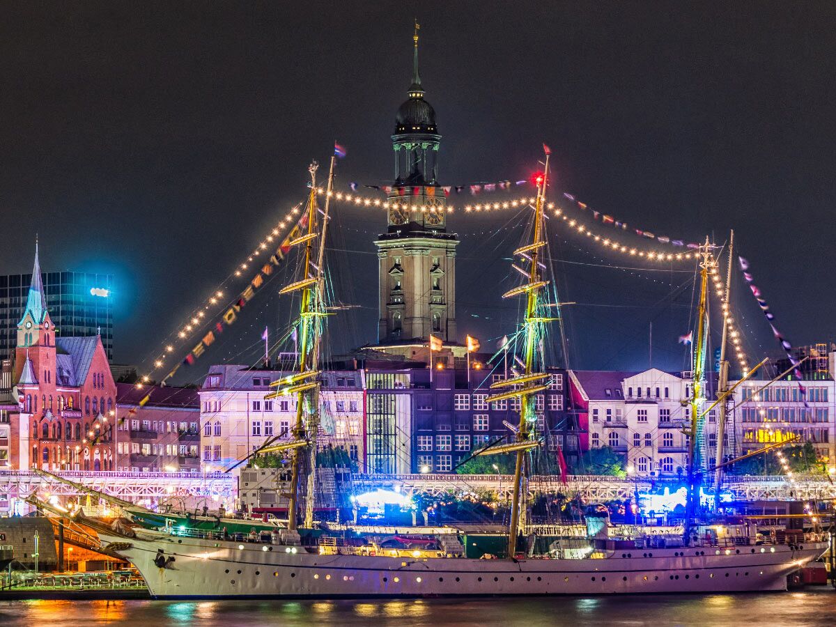 Großes, festlich beleuchtetes Segelschiff nachts im Hamburger Hafen mit Lichterketten und historischem Kirchturm im Hintergrund.