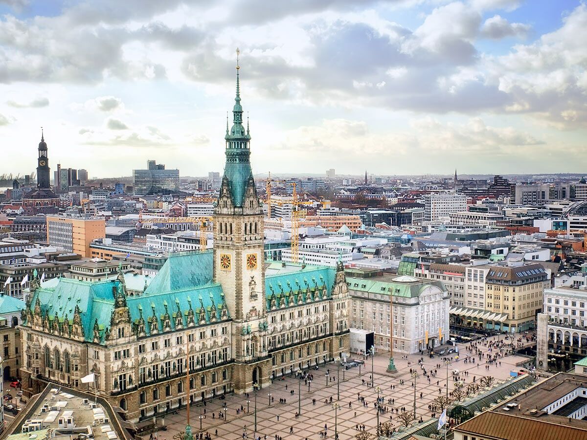 Luftbild vom Hamburger Rathaus mit Rathausmarkt, Menschen, historischen Gebäuden und Skyline bei bewölktem Himmel.