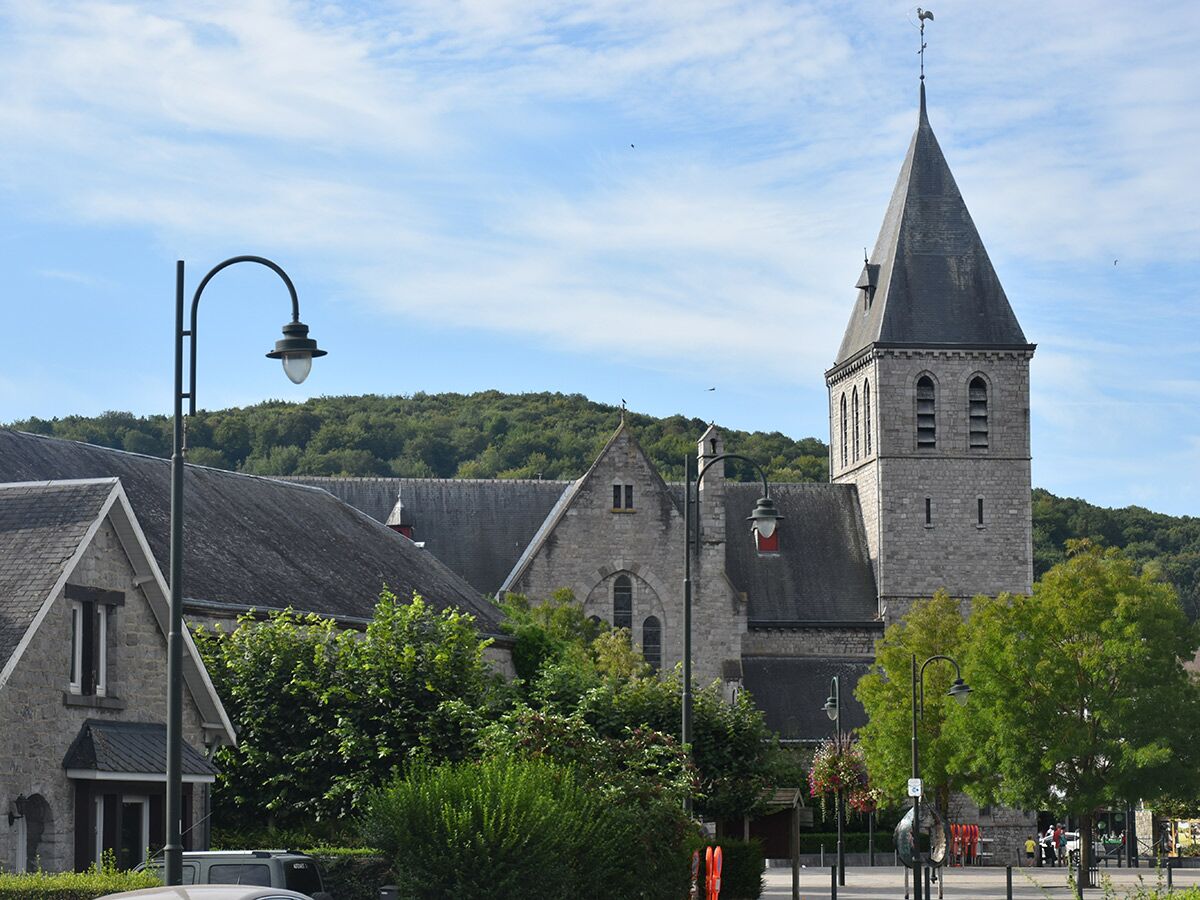 Historische Steinkirche mit spitzem Kirchturm in einem europäischen Dorf, umgeben von grünen Bäumen und Laternen unter blauem Himmel.