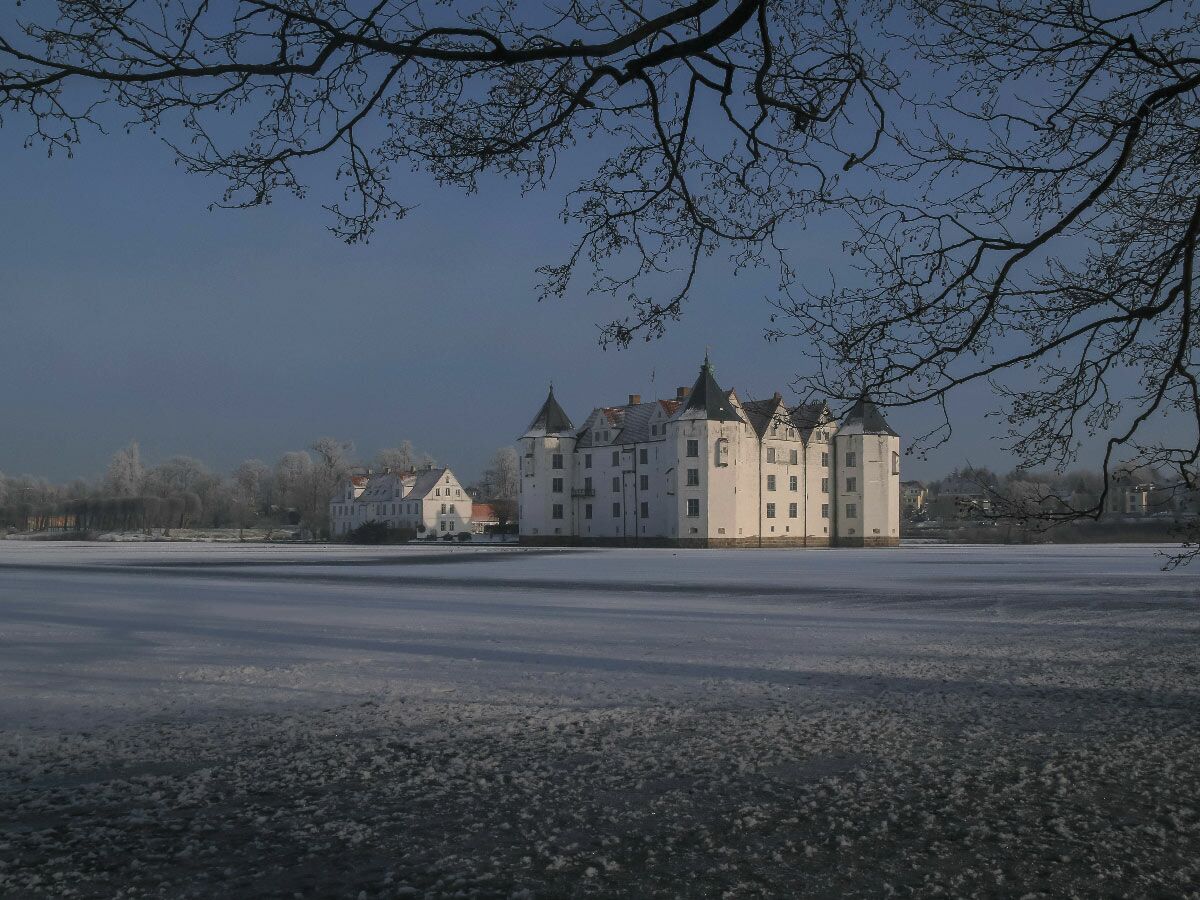 Weißes Wasserschloss mit dunklen Dächern an gefrorenem See im Winter mit verschneiten Bäumen unter blauem Himmel.