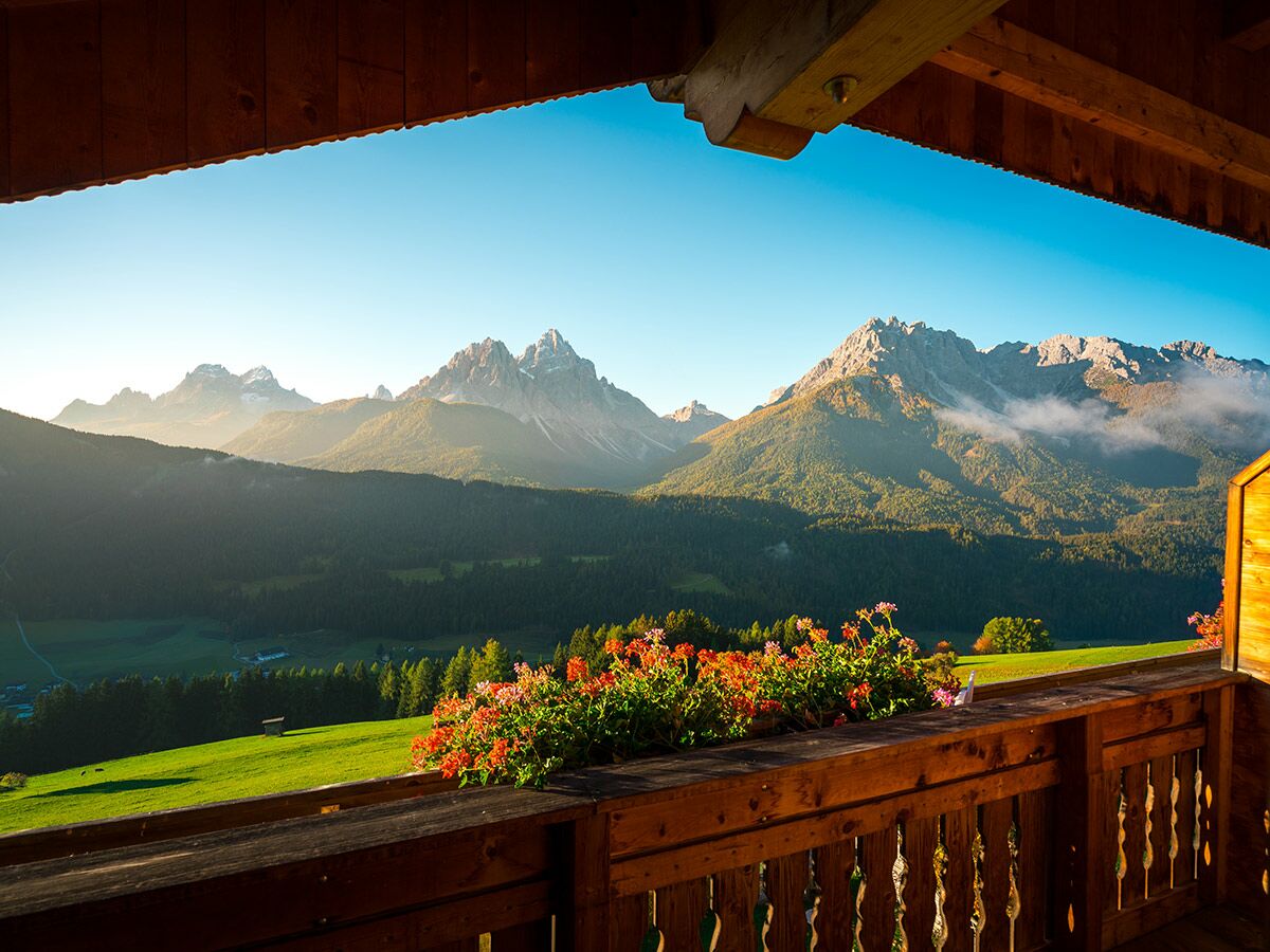 Blick vom hölzernen Balkon mit bunten Blumenkästen auf ein sonniges Bergpanorama der Alpen mit grünen Tälern und Wäldern.