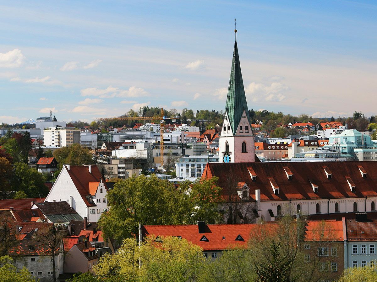 Panoramaansicht einer charmanten deutschen Stadt mit einer hohen Kirchturmspitze, roten Dächern, Bäumen und Bergen im Hintergrund.