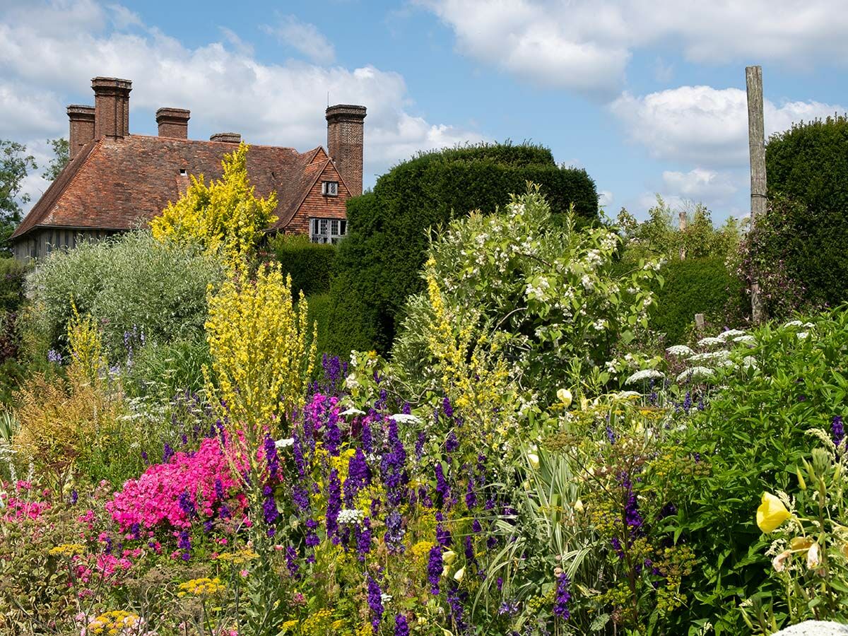 Vibranter, bunter Garten mit gelben, lila, pinken Blumen und historischem Haus mit Ziegeldach, Schornsteinen unter blauem Himmel.
