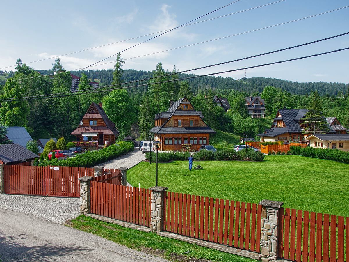 Traditionelle Holzhäuser in einem polnischen Bergdorf mit grünen Hängen, Wald und blauem Himmel. Ein Mann mäht den Rasen vor einem roten Holzzaun.