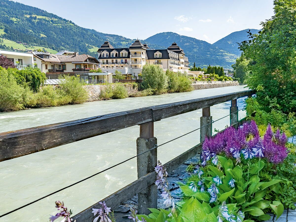 Malerische Flusslandschaft mit historischem Alpenhotel und modernen Häusern vor grünen Bergen, betont durch blühende Blumen im Vordergrund.