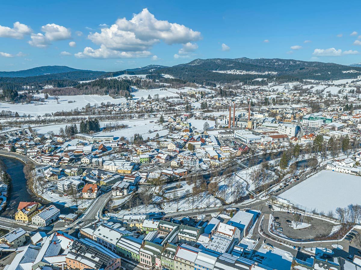 Luftaufnahme einer idyllischen, verschneiten Stadt in einem Tal, umgeben von schneebedeckten Bergen und Wäldern unter blauem Himmel.