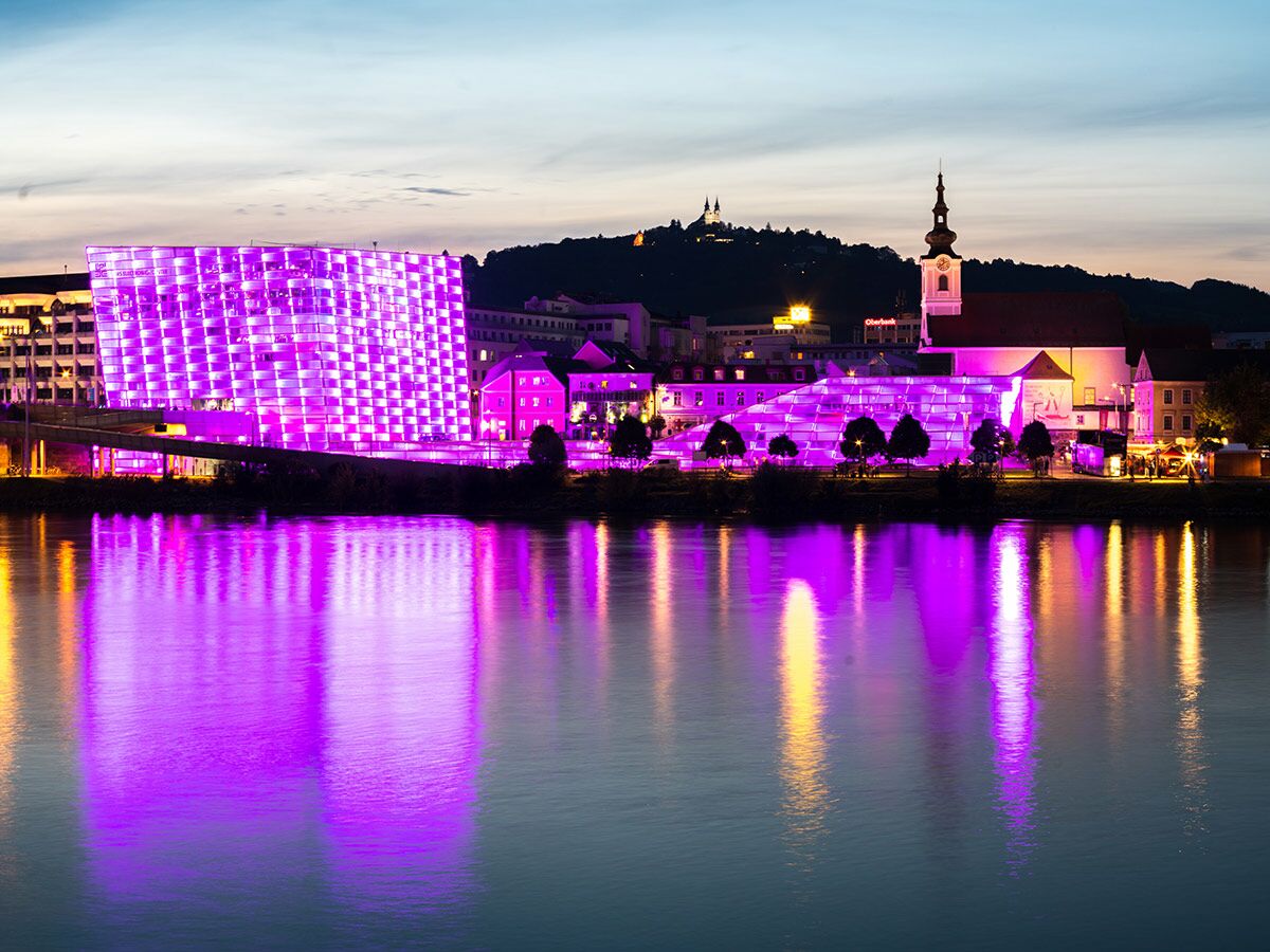 Linz: Das lila leuchtende Ars Electronica Center und eine Kirche bei Nacht, gespiegelt in der Donau vor dem Pöstlingberg.