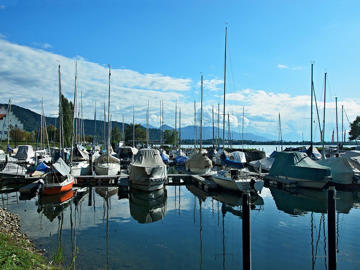 Viele Segelboote liegen im Hafen eines Sees, umgeben von Bergen und blauem Himmel, ihre Masten spiegeln sich im Wasser.