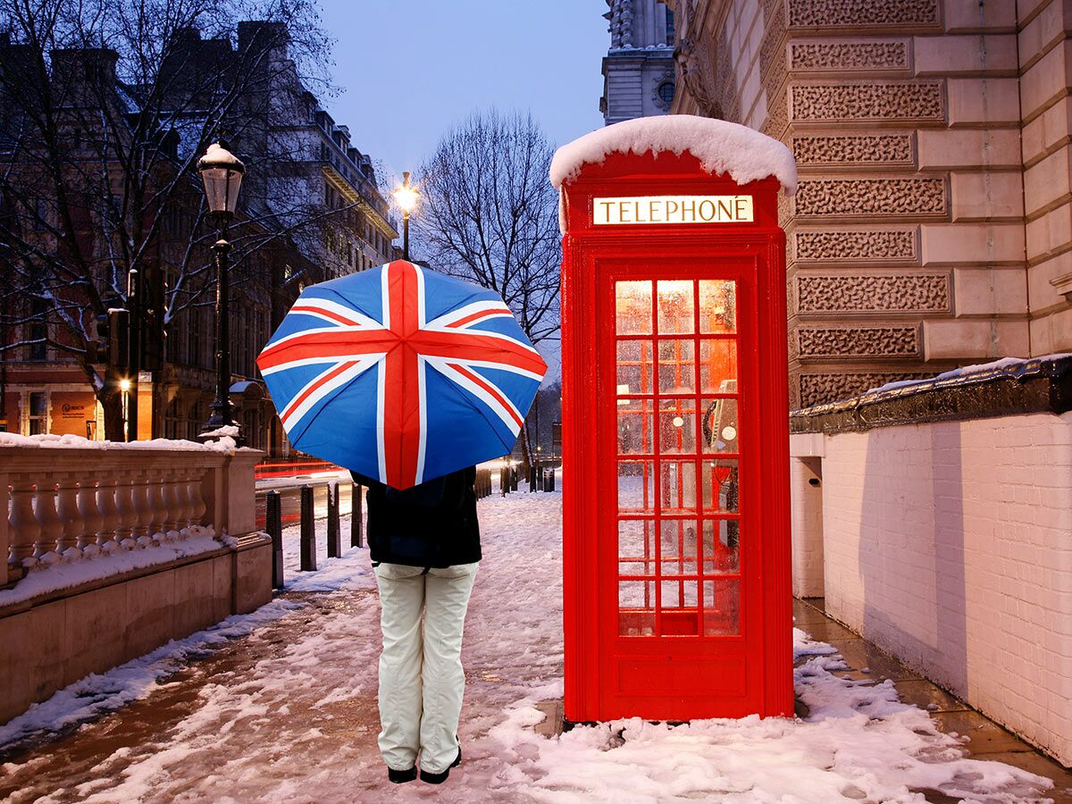Person mit Union Jack Regenschirm steht neben klassischer roter Londoner Telefonzelle im Schnee einer winterlichen Straße.