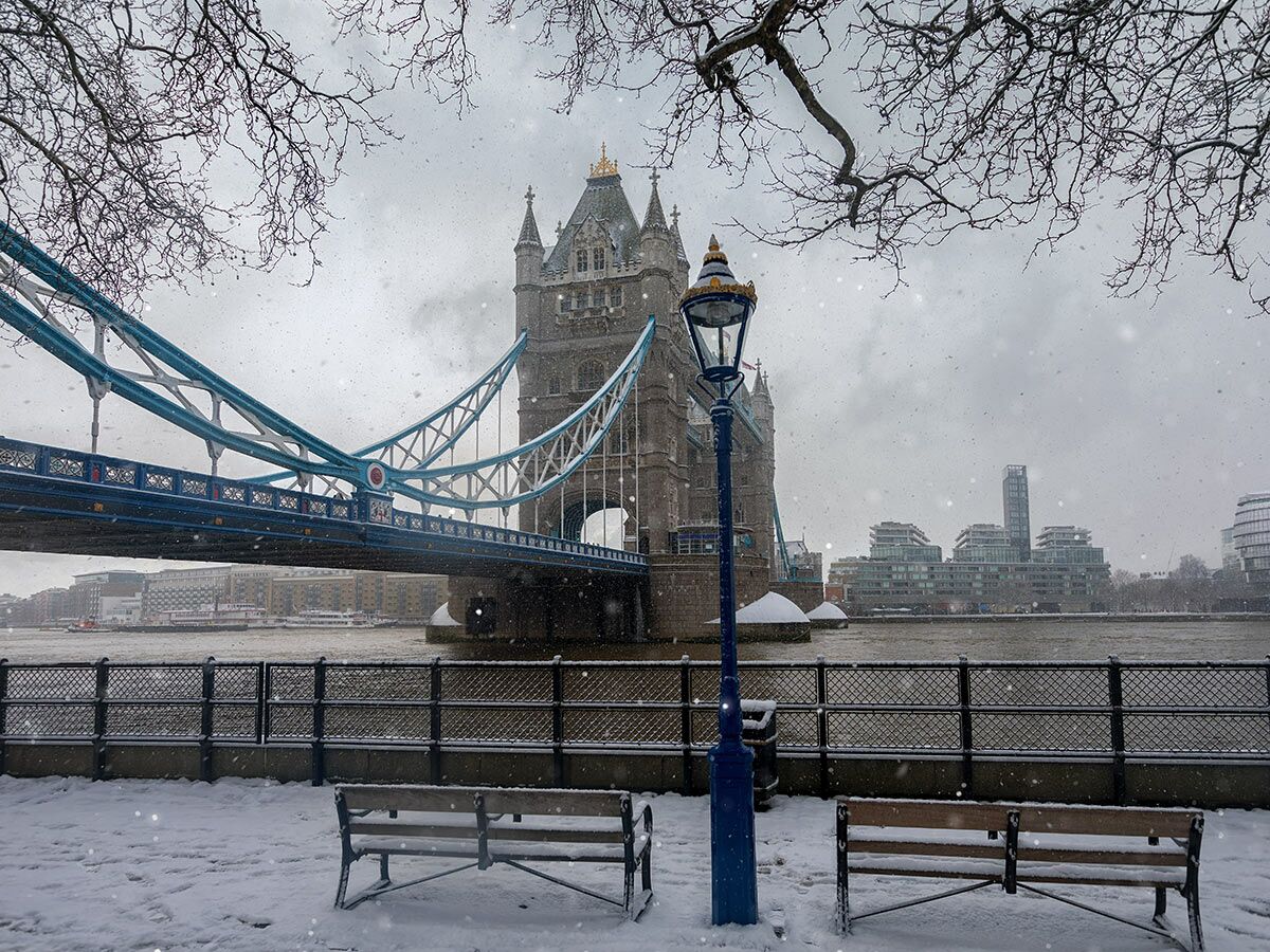 London Tower Bridge im Schnee mit Lampe, Bänken und Bäumen am Themseufer.