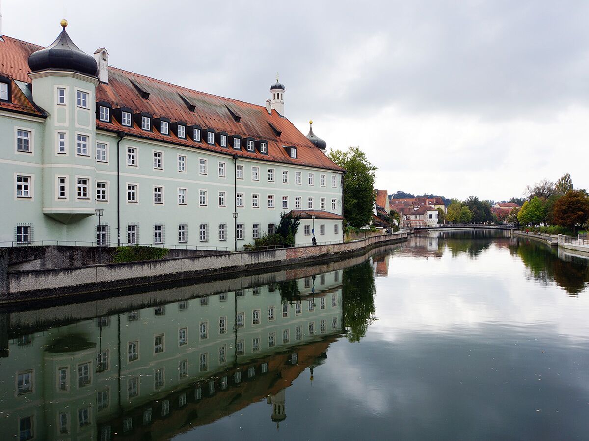 Langes, hellgrünes Gebäude mit rotem Dach und Kuppelturm am Fluss, dessen Spiegelung im ruhigen Wasser sichtbar ist.