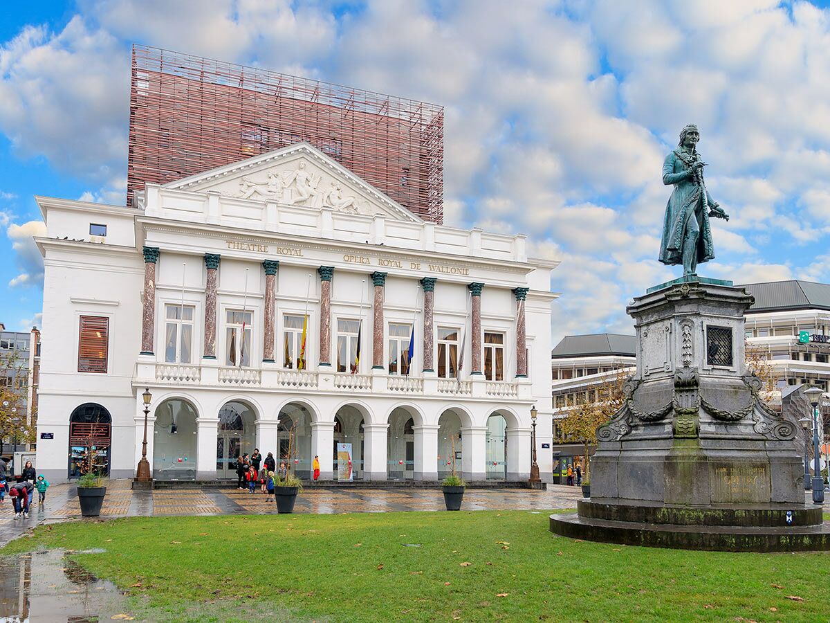 Klassisches weißes Gebäude der Königlichen Oper Wallonie in Lüttich mit Säulen und belgischen Flaggen, davor ein Bronzedenkmal.