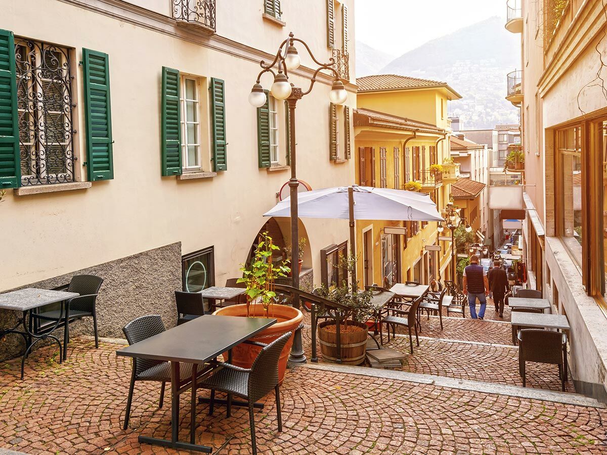 Blick auf eine charmante italienische Altstadtgasse mit Kopfsteinpflaster, Cafétischen und grünen Fensterläden an sonnigem Tag.