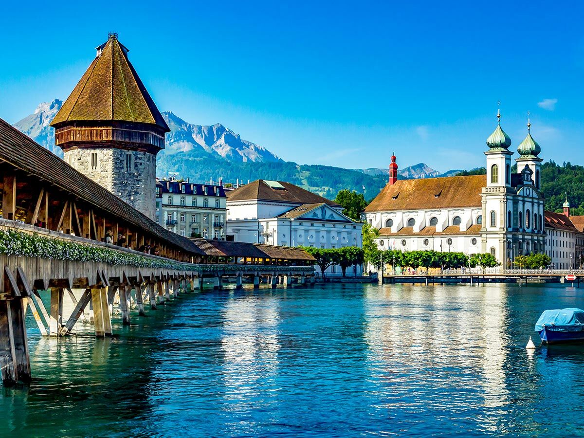 Historische Kapellbrücke mit Wasserturm, Jesuitenkirche und Fluss Reuss in Luzern, Schweiz, vor alpiner Bergkulisse.