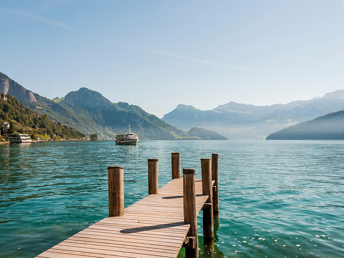 Holzsteg ragt in blauen Bergsee mit Schiff, umgeben von grünen Bergen und klarem Himmel an einem sonnigen Tag.